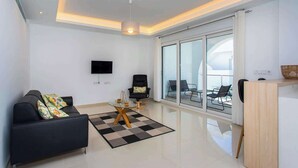 Un salon moderne avec un canapé, une table basse et un téléviseur. Une grande fenêtre offre une vue sur l’extérieur.