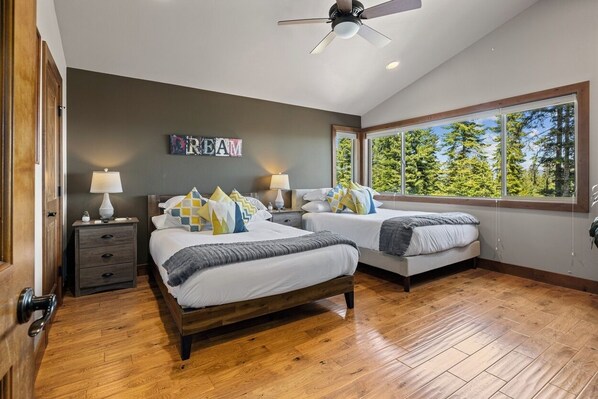A bedroom with two beds, wooden flooring, a ceiling fan, and a large window with a view of trees.
