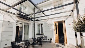 A covered outdoor seating area with tables and chairs, a glass ceiling, and a wooden door leading inside. - Hotel Flaneur (Belgrade)