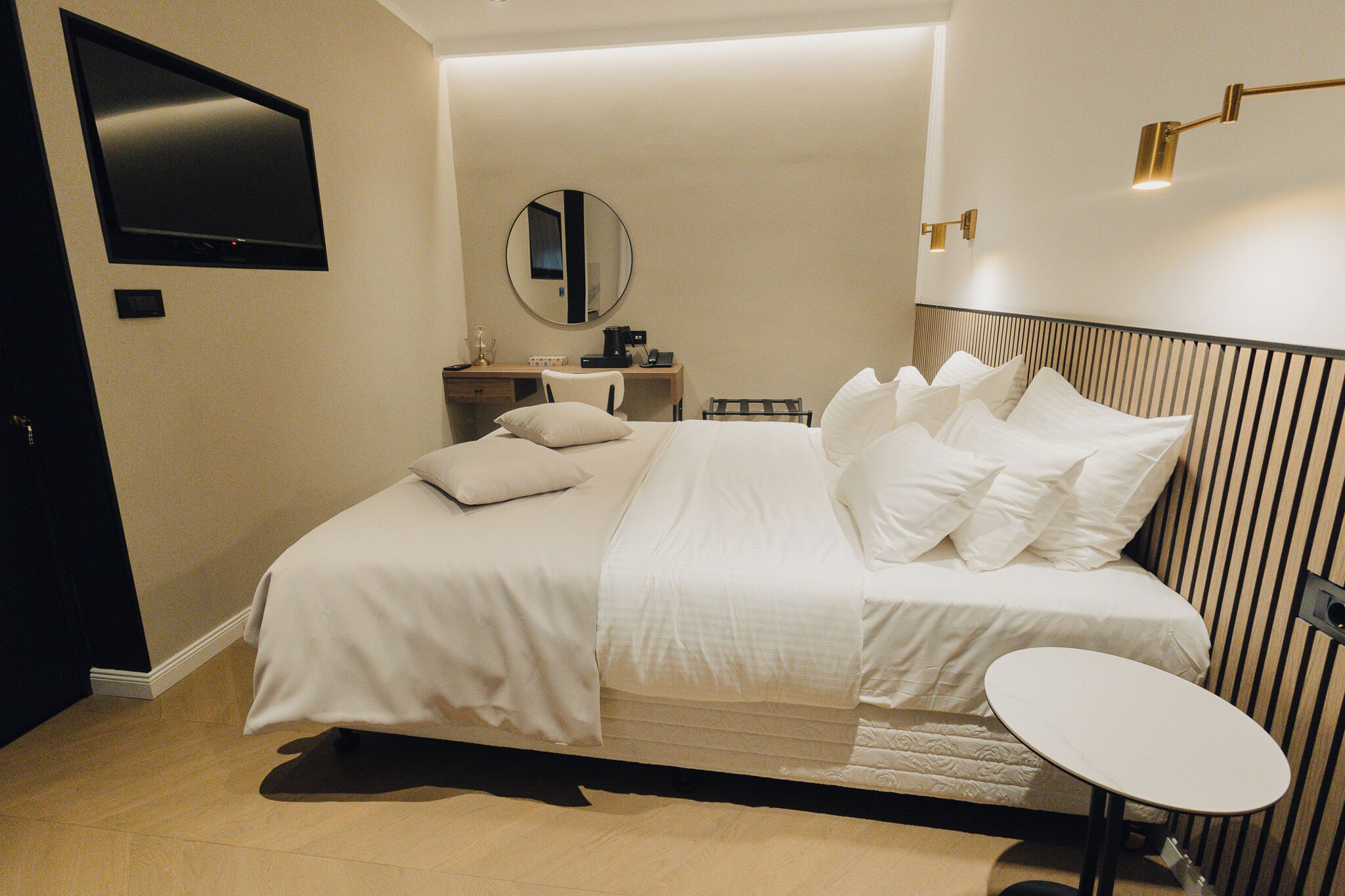 A modern hotel room with a large bed, a small table, a TV, a mirror, and a bedside lamp.
