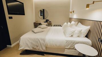 A modern hotel room with a large bed, a small table, a TV, a mirror, and a bedside lamp.