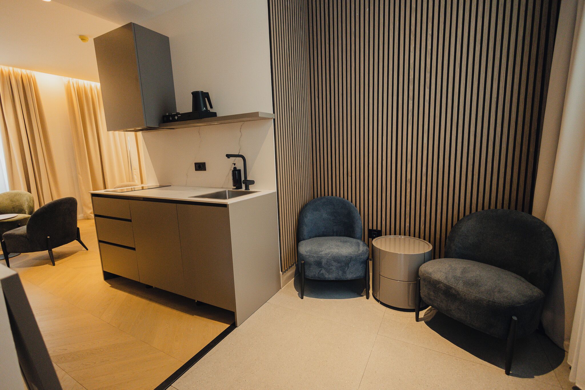 A modern kitchen with a built-in sink, a coffee machine, and a small trash bin. There are two dark chairs and a small table in the room.