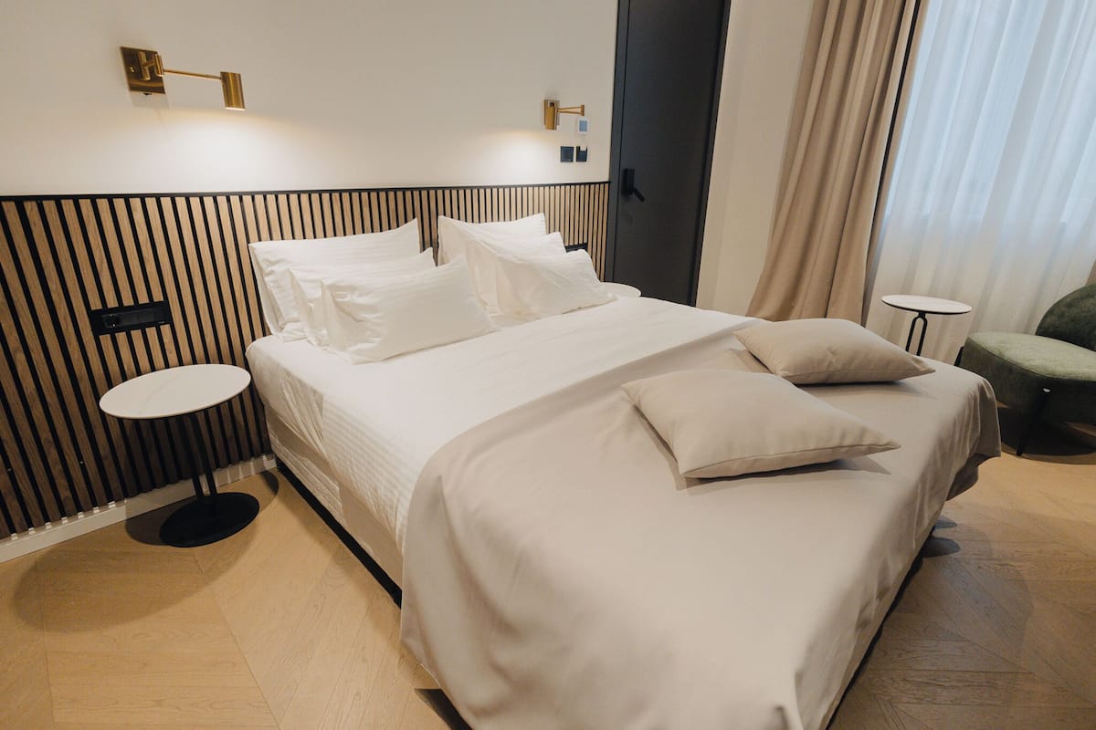 Un lit bien fait, avec du linge de lit blanc et des oreillers, une table d’appoint ronde, une petite table ronde et un fauteuil vert dans une chambre d’hôtel moderne.
