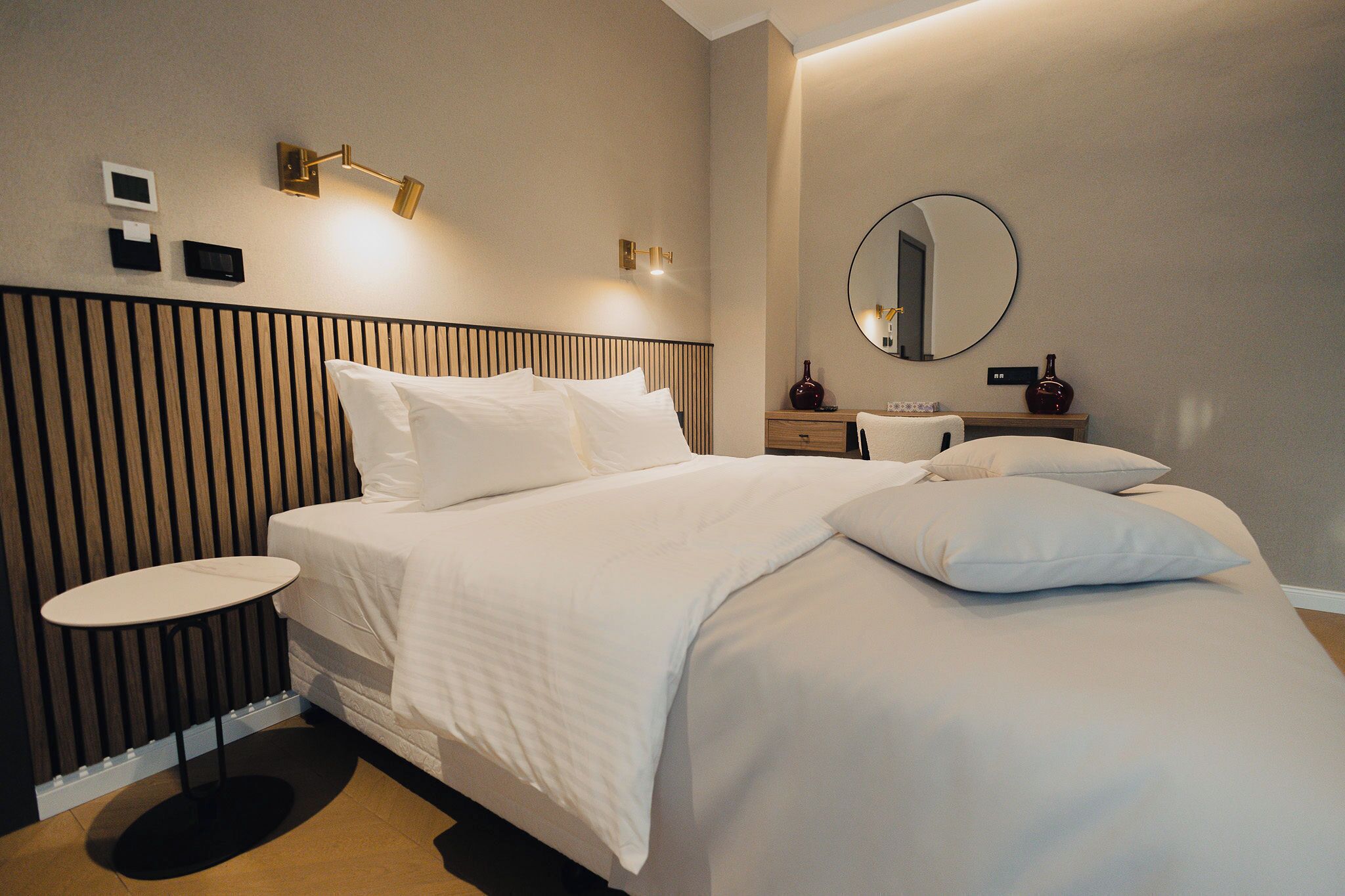 A modern hotel room with a large bed, bedside tables, a round mirror, and wall-mounted lighting.