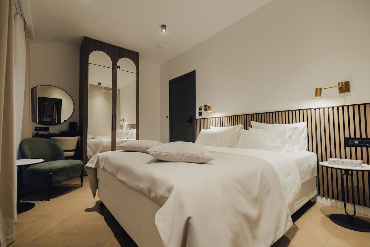 Une chambre d’hôtel avec un grand lit, une chaise, une petite table, un miroir et une table de chevet avec une lampe.