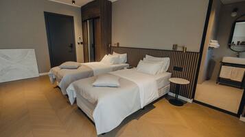 Une chambre d’hôtel moderne avec deux lits simples, une petite table ronde, un banc intégré et une salle de bain avec un miroir et un lavabo.