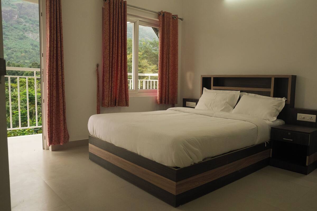 Une chambre à coucher avec un lit, des tables de chevet, une tête de lit en bois, une fenêtre avec des rideaux et une vue sur la verdure à l’extérieur.