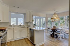 Uma cozinha moderna com armários brancos, máquina de lavar louça embutida e uma ilha grande com pia. Há uma área de jantar com cadeiras brancas e um lustre.