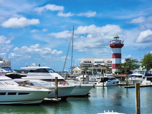 Uma marina com vários iates atracados, um farol ao fundo e um edifício com área externa para refeições.