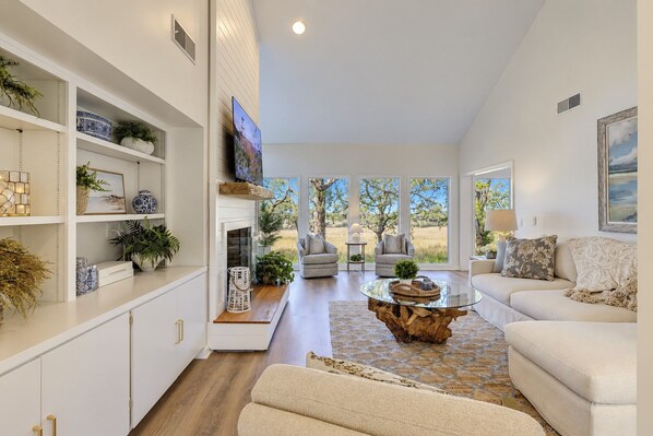 A spacious living room with a fireplace, comfortable seating, and large windows offering a view of the outdoors.