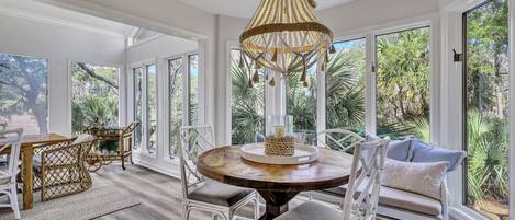 A spacious room with a round wooden table, white wicker chairs, and a large window with a view of greenery.