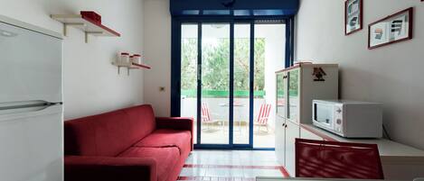 A compact living space with a red sofa, a white refrigerator, a microwave, and a dining area with a table and chairs.