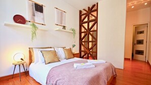 Een slaapkamer met houten vloer, een bed met een wit-bruine dekbedovertrek, een nachtkastje met een lamp en een hangklok.