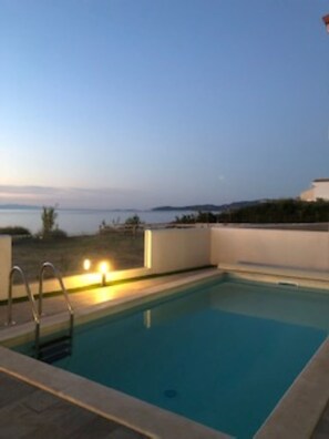 Una piscina rectangular con una escalera metálica, rodeada por un muro blanco bajo, con vistas al mar y a la costa lejana bajo un cielo despejado.