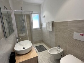 Un baño moderno con un lavamanos ovalado grande, un lavamanos más pequeño, un box de ducha de vidrio y un perchero en la pared.