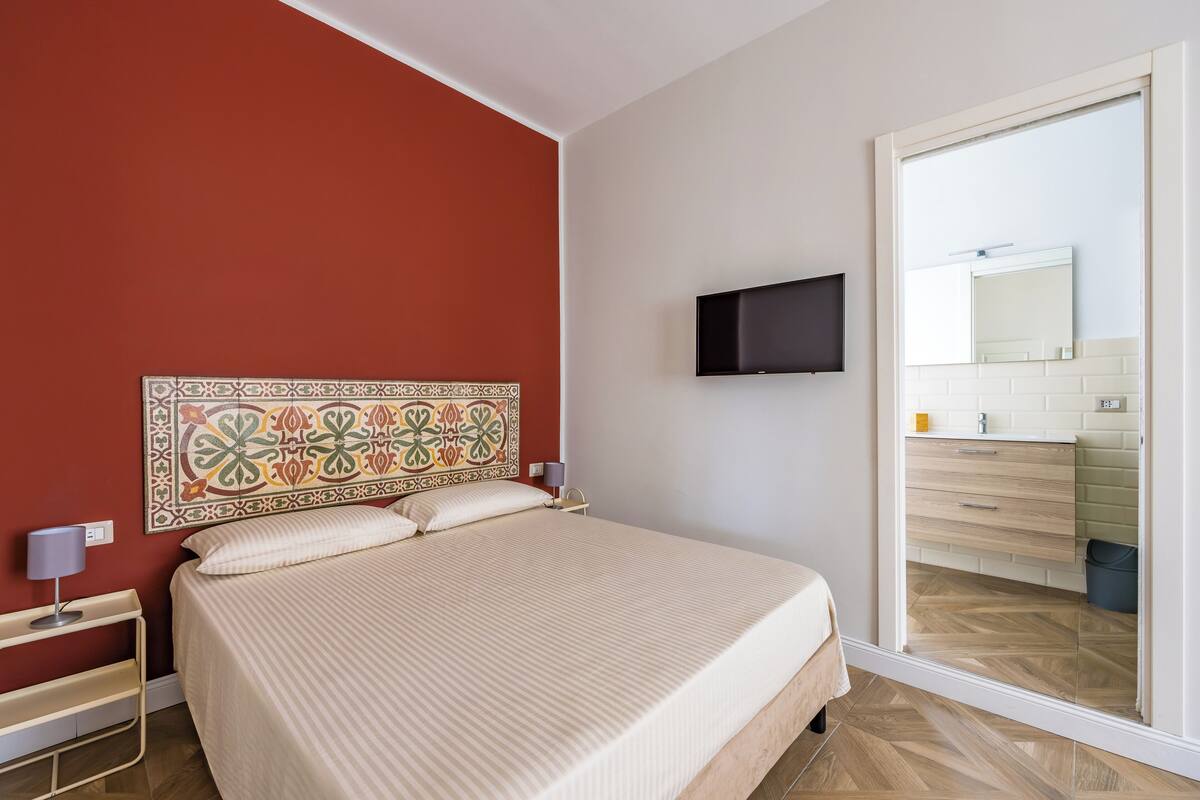Hotellihuone, jossa on sänky, seinälle asennettu televisio, avoimesta ovesta näkyvä kylpyhuone ja kuvioitu pääty.