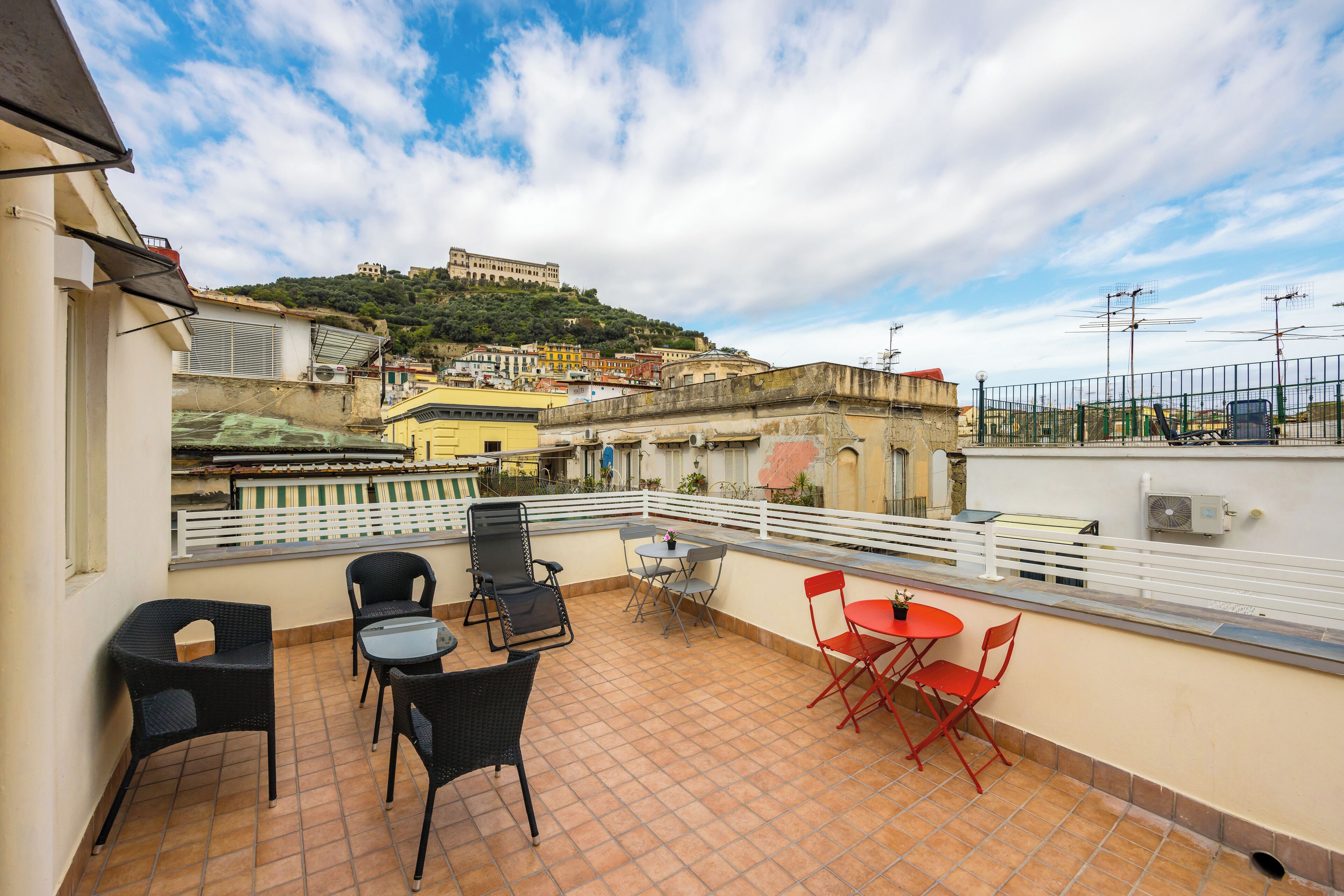 Une terrasse avec des sièges extérieurs, une vue sur une colline parsemée de bâtiments et une rambarde blanche.