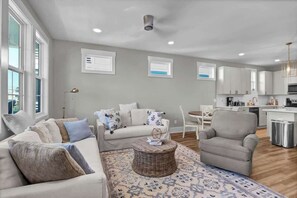 A modern living room with a sofa, armchair, and a small round table. There is a kitchen area with cabinets and a microwave. - Shenanigans on the Sea (SOS) (Carolina Beach)