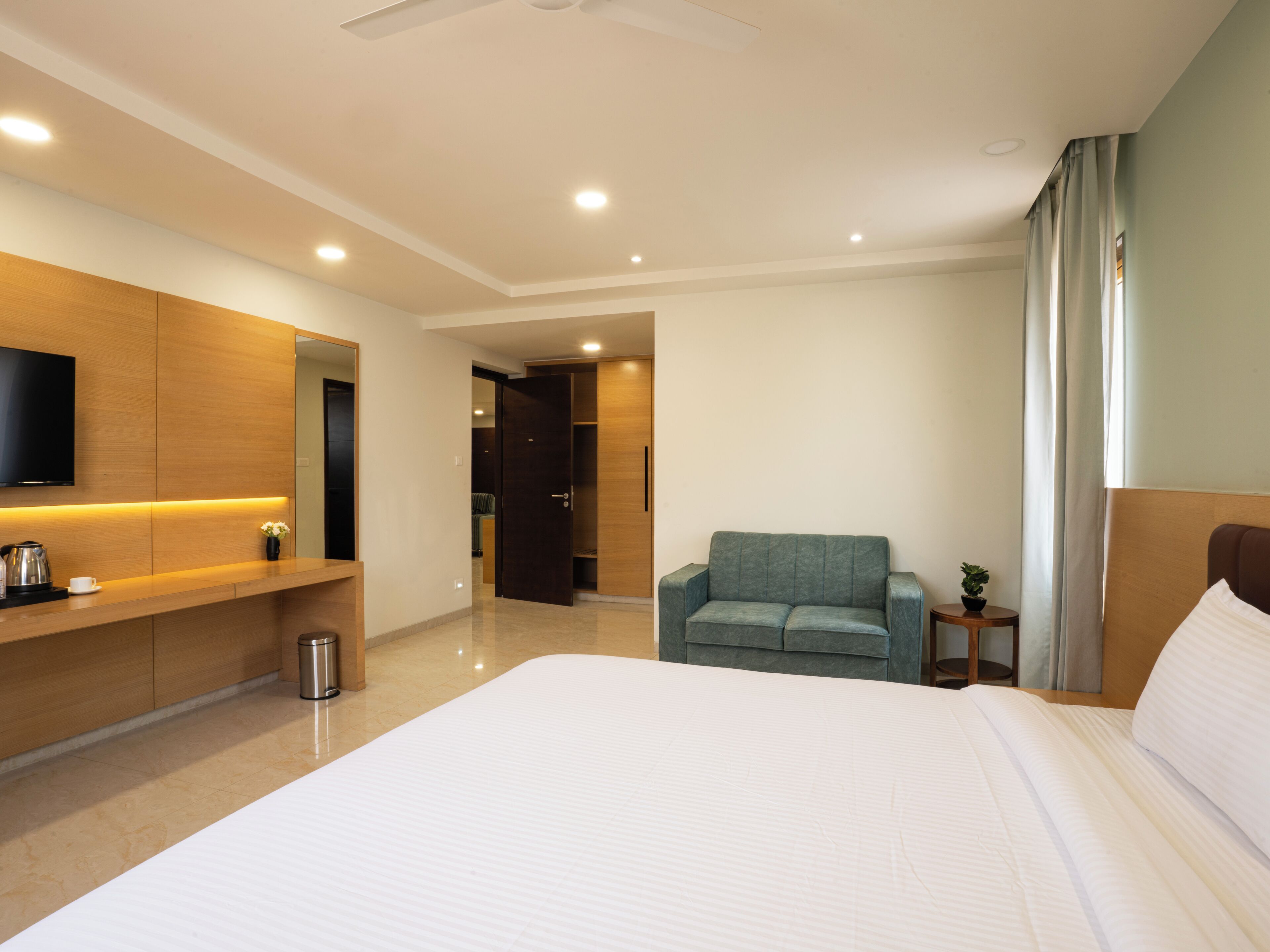 A modern hotel room with a large bed, a seating area with a sofa, a TV mounted on the wall, and a small table with a plant.