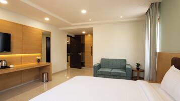 A modern hotel room with a large bed, a seating area with a sofa, a TV mounted on the wall, and a small table with a plant.