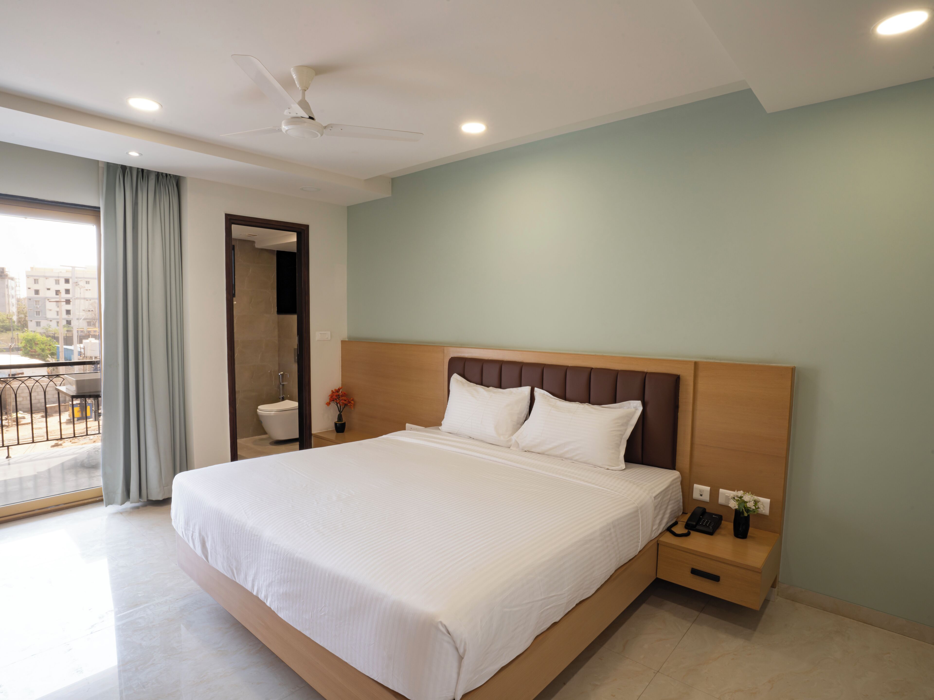 Una habitación de hotel moderna con una cama grande, un cabecero de madera, una mesita de noche con un teléfono y un jarrón, y vistas a la ciudad desde un balcón.