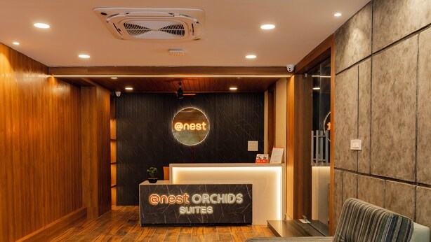 A reception area with a wooden floor, a reception desk labeled "Nest Orchids Suites," and a seating area with a striped cushion.