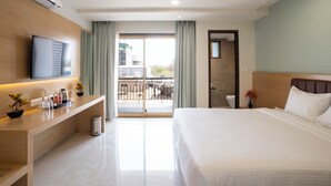 A hotel room with a bed, a TV, a desk with a trash bin, and a balcony with a view of buildings.
