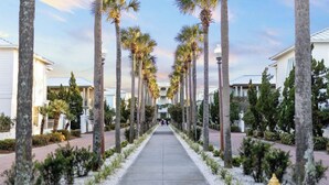 A pathway lined with palm trees leading to a building with a white facade. - Tranquil Shores | Walk to Beach | Largest 30A Pool | Sleeps 12 | 6 Bikes (Panama City Beach)