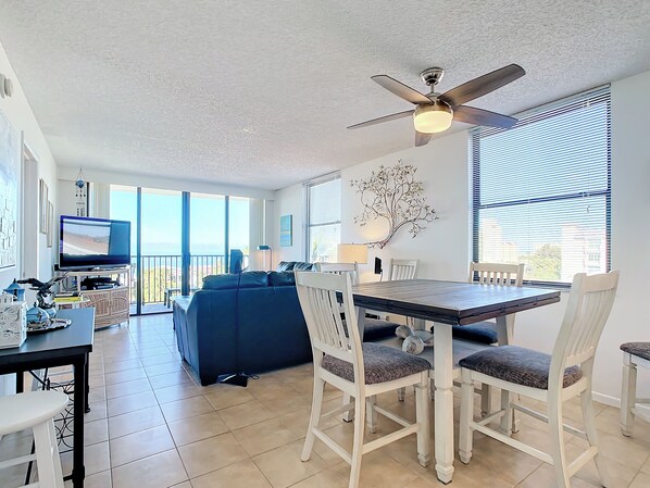 A modern living room with a dining area, a ceiling fan, large windows, and a TV. - Quiet Waters Condominium 4E-Perfect beach getaway! (Indian Shores)