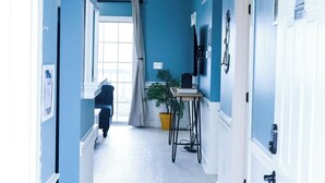 A hallway with blue walls, white doors, and a ceiling light fixture. There is a window with curtains, a plant on a table, and a door with a sign.
