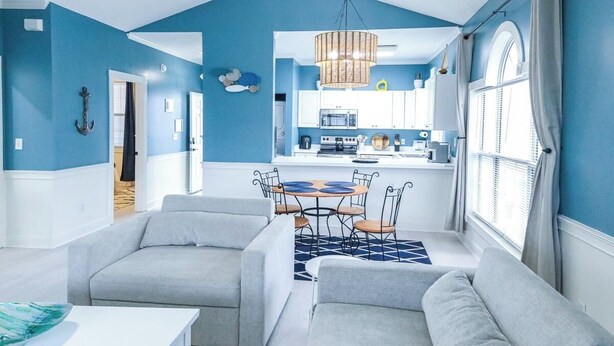 A modern living room with a dining area, a kitchen in the background, and a large window with curtains.