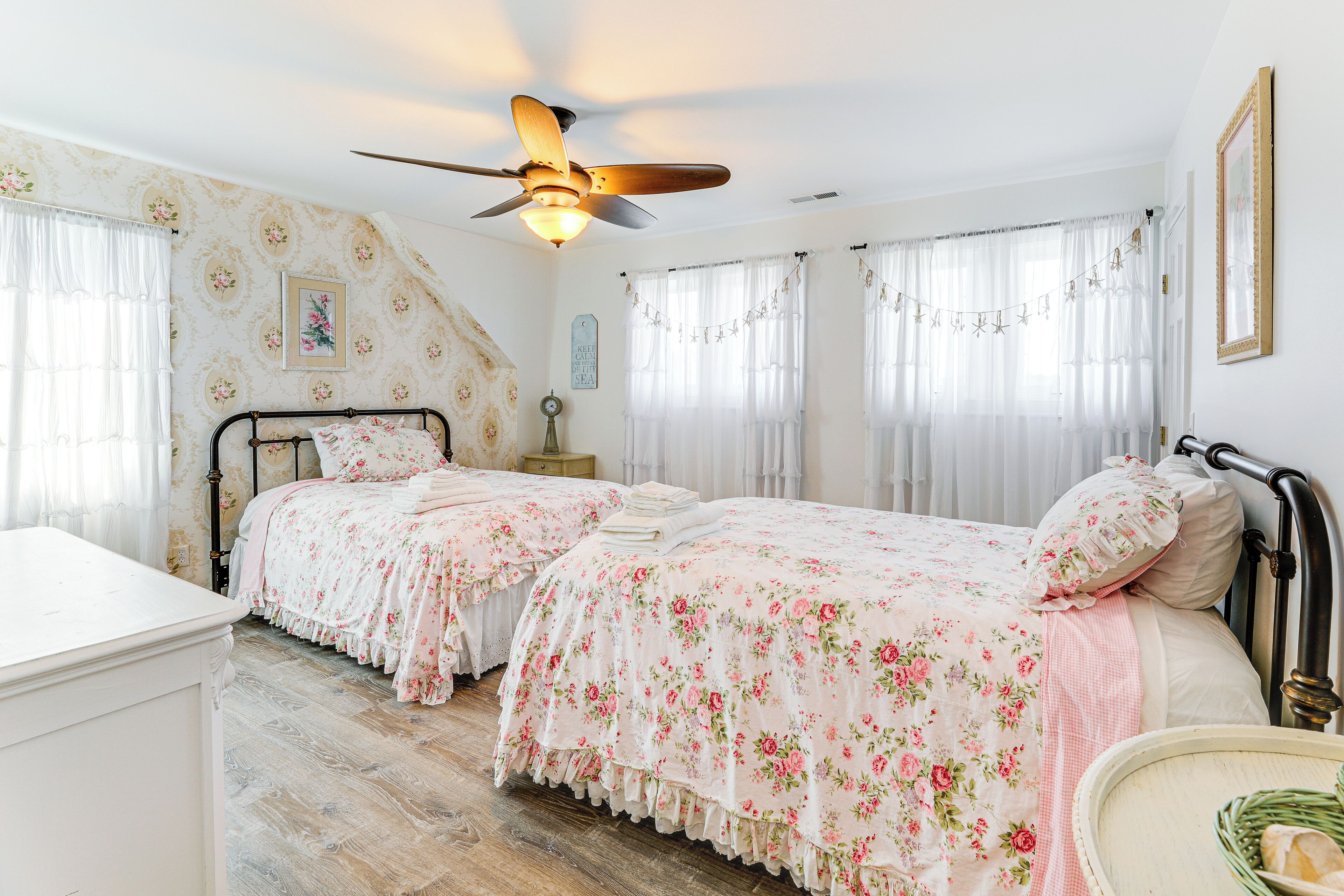 A bedroom with two beds, floral bedding, a ceiling fan, and a bedside table with a lamp.