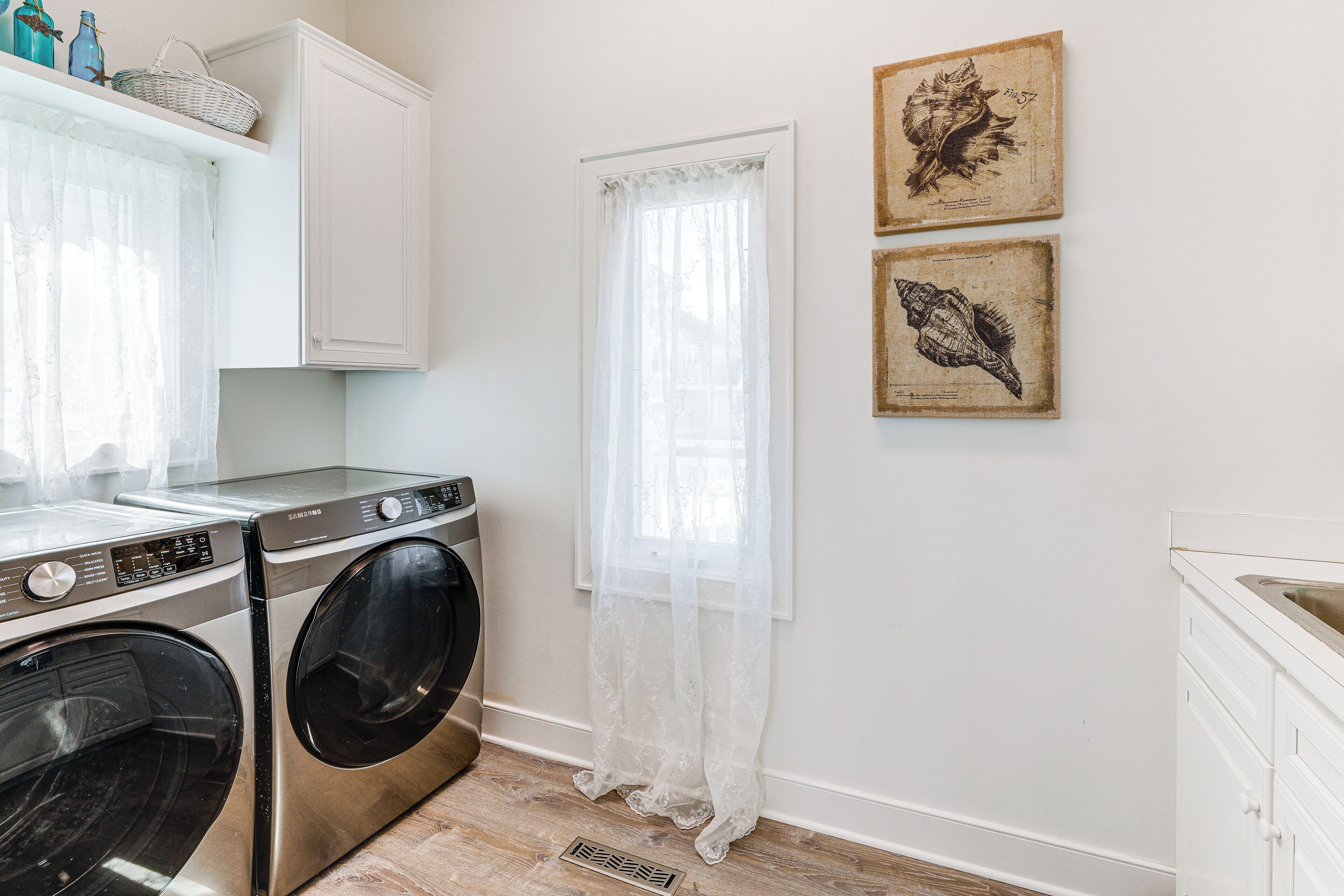 A laundry room with a washing machine and dryer, a window with sheer curtains, and two framed pictures on the wall.