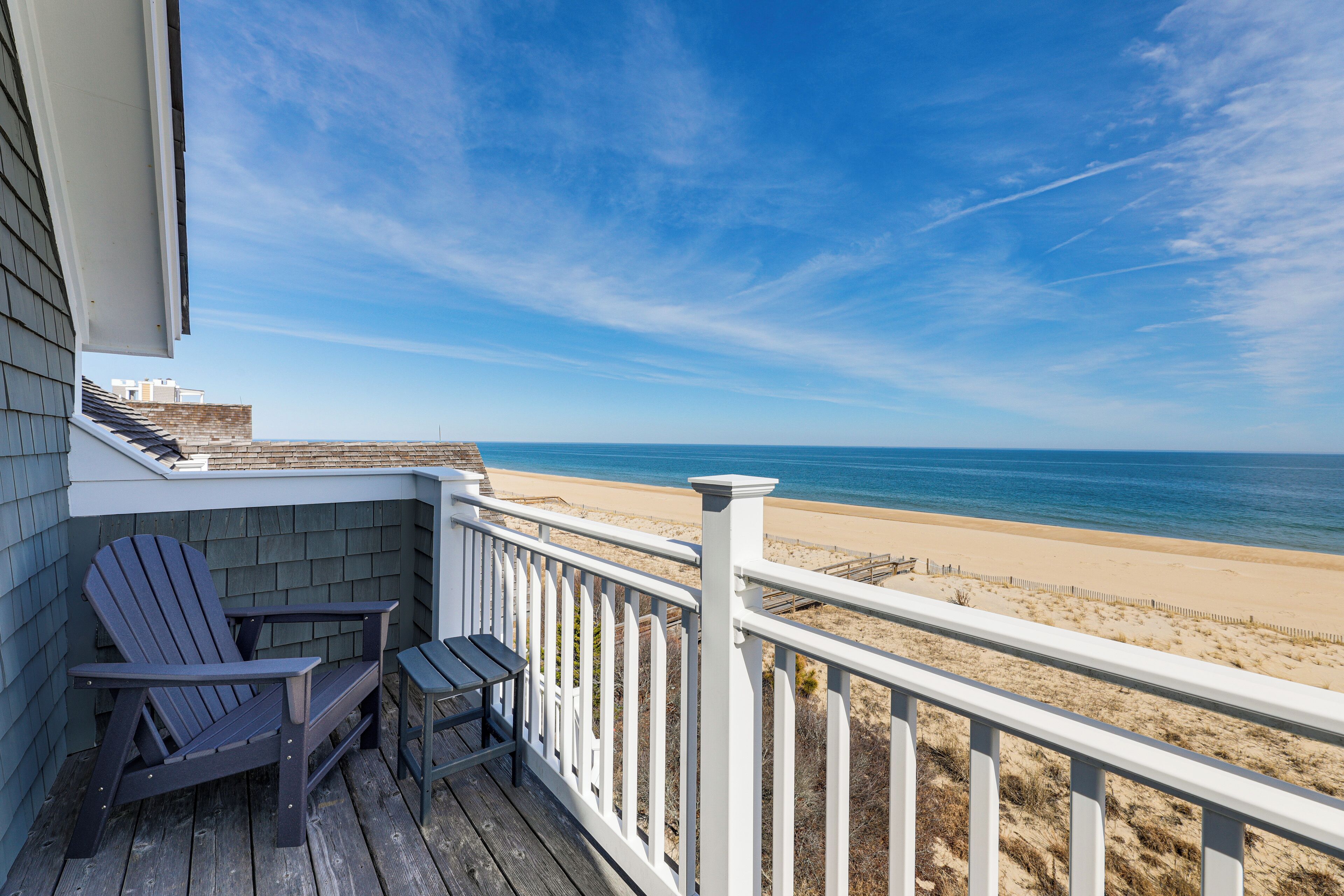 Eine Holzterrasse mit Adirondack-Stühlen und einem Tisch mit Blick auf einen Strand.