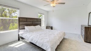 A bedroom with a bed, a ceiling fan, a nightstand, a dresser, and large windows.