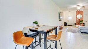 A modern dining area with a wooden table and four chairs, a small plant on the table, and a living area with a sofa and a plant in the background.