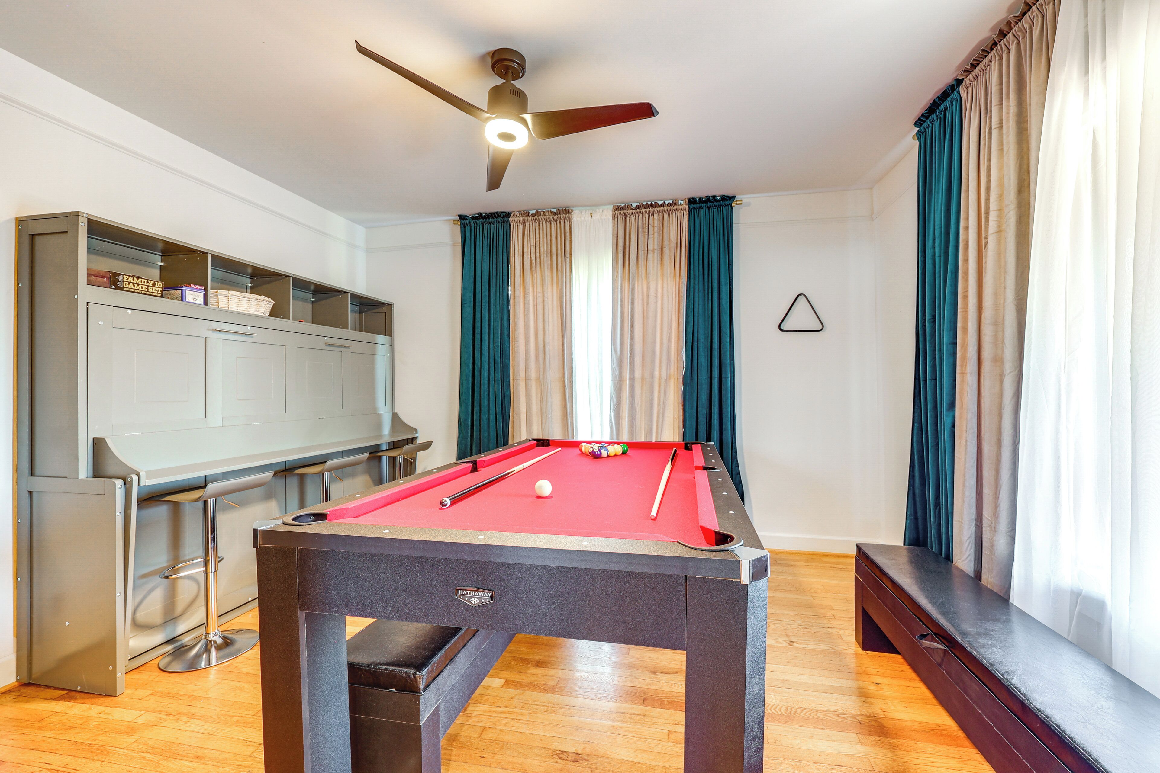 A pool table with a red felt surface, a ceiling fan, and a bar area with stools.