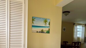 A room with a white door, a painting of a beach scene, a dining area with wooden furniture, and a tiled floor.