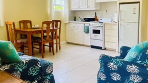 A compact kitchen with white cabinets, a microwave, a stove, and a refrigerator. There is a dining table with chairs and a blue sofa with a coral pattern.