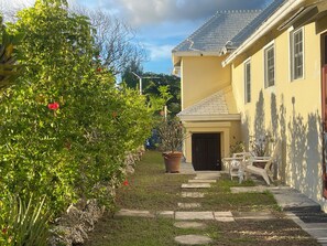 Un bâtiment jaune avec un toit en tuiles, un chemin pavé menant à l’entrée et un jardin agrémenté d’arbustes verts et de fleurs rouges.