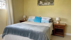 A neatly made bed with a grey and white duvet, two bedside tables with lamps, a painting on the wall, and a window with white curtains.