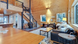 A spacious living area with a wooden ceiling, a staircase, a fireplace, and a large window. - Hot Tub & Sauna: Mountain Living at Hyak Cabin! (Snoqualmie Pass)