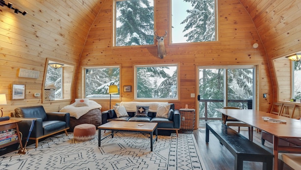 A cozy log cabin interior with a wooden ceiling, large windows, a sofa, a coffee table, and a dining table. - Hot Tub & Sauna: Mountain Living at Hyak Cabin! (Snoqualmie Pass)