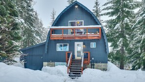 A two-story cabin with a wooden staircase leading to the entrance, surrounded by snow-covered trees. - Hot Tub & Sauna: Mountain Living at Hyak Cabin! (Snoqualmie Pass)