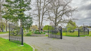 A gated entrance with a decorative metal gate, a gravel driveway, and a grassy area with trees. - 9 Mi to Spanaway Park: Home w/ Indoor Hot Tub (Spanaway)