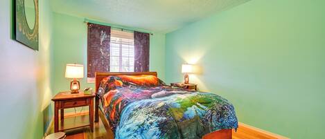 A bedroom with a wooden bed, two bedside tables with lamps, a nightstand, a trash bin, and a window with curtains.