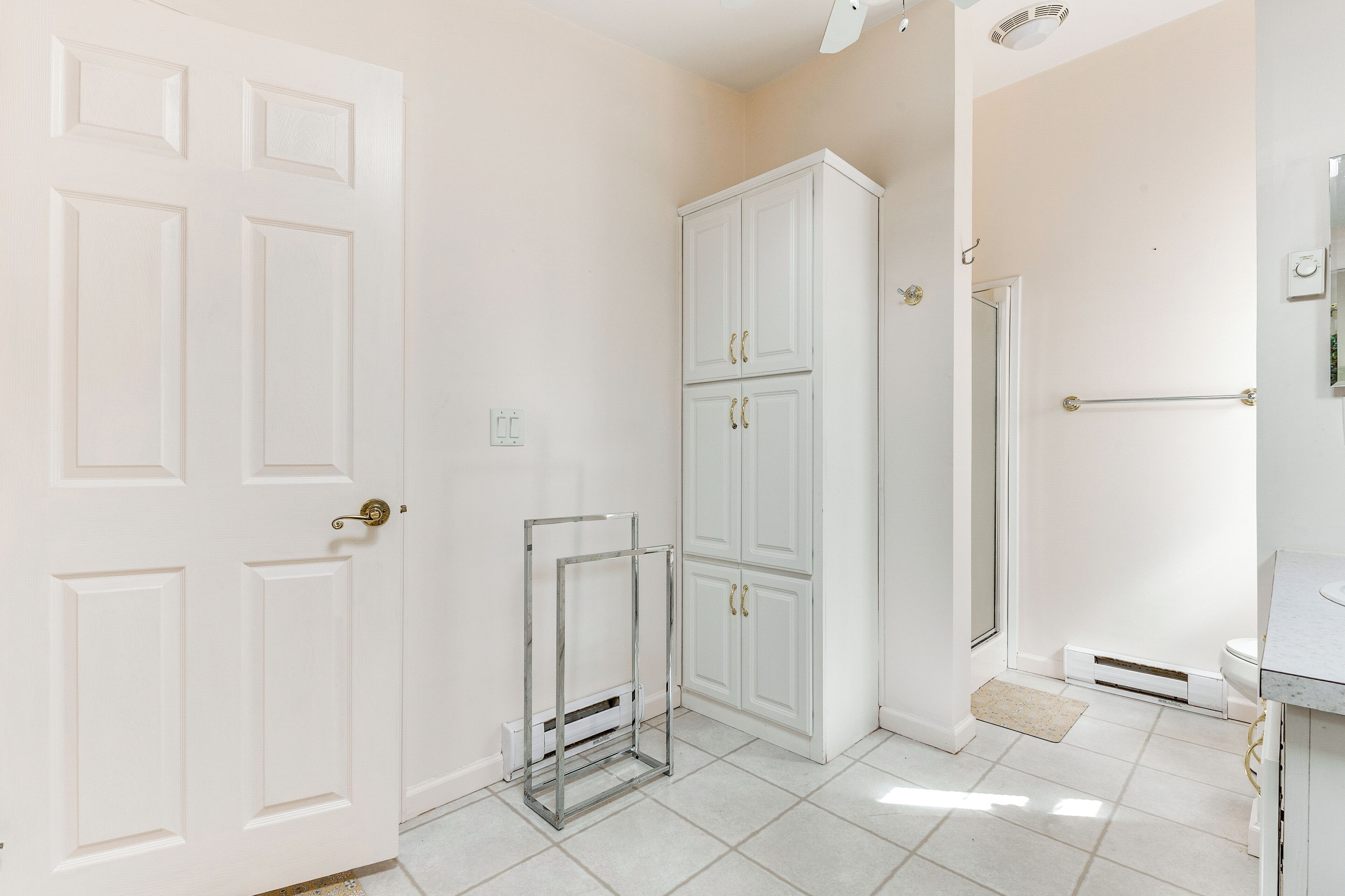 A bathroom with white tiled floors, a white door, a white cabinet with two drawers, a glass shelf unit, and a white sink with a countertop.