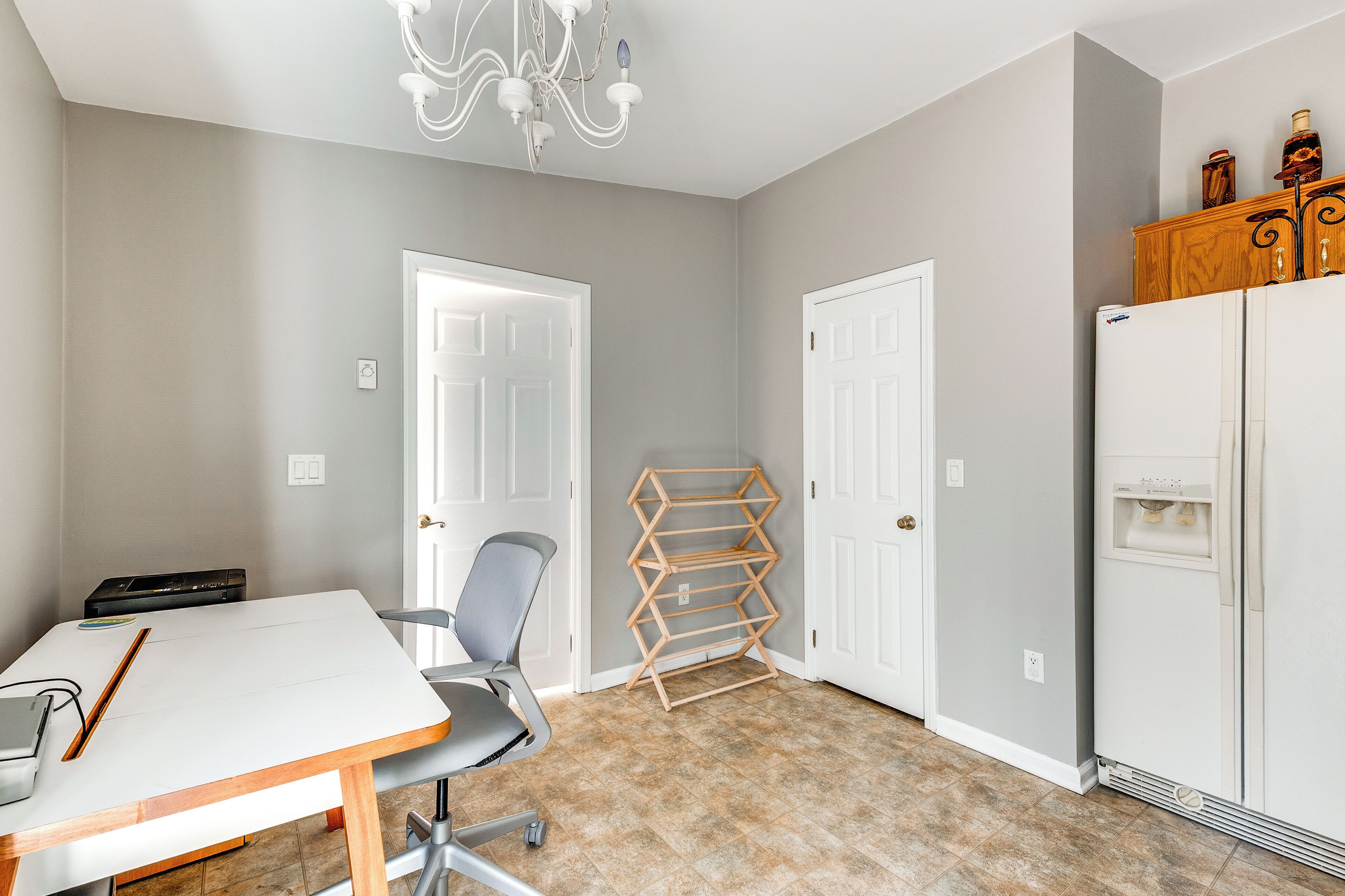Une pièce avec une table blanche, une chaise grise, un étendoir en bois et un réfrigérateur blanc.