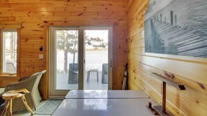 Interior de una cabaña de madera con una ventana grande, una mesa de comedor, sillas de mimbre y vista a un muelle y al agua.