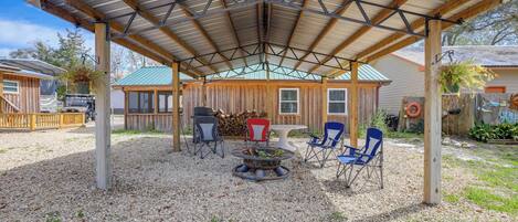 A covered outdoor seating area with a metal roof, surrounded by gravel, featuring a fire pit, and furnished with multiple chairs.